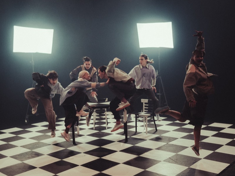 Six dancers suspended in mid-air around a cluster of white wooden chairs, backlit by two large panels.
