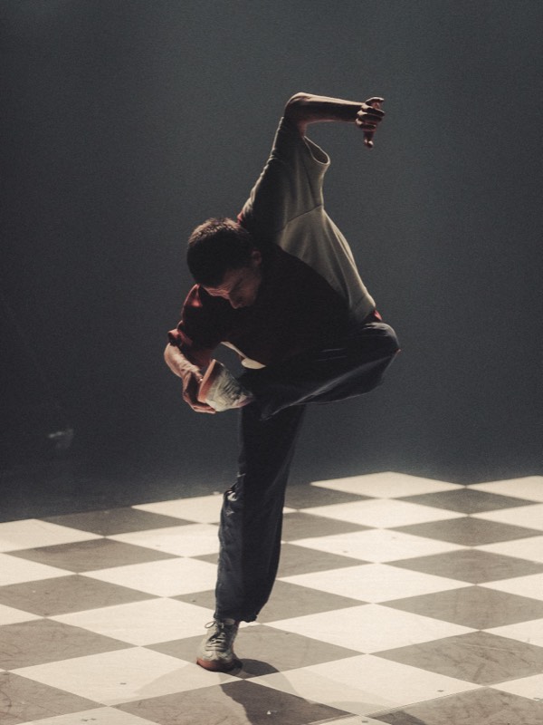 A single dancer folded deep over one leg in hard backlight, in a spliced olive tee and loose navy trousers.