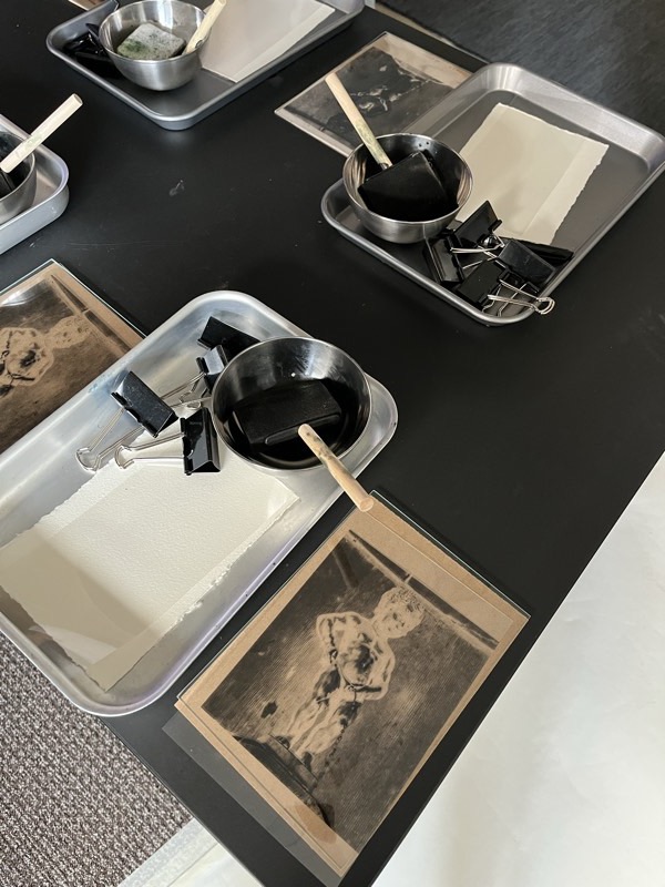 Prepared workshop stations on a black table: steel trays of deckle-edged paper, cups of dye and wooden stirrers stained yellow-green.