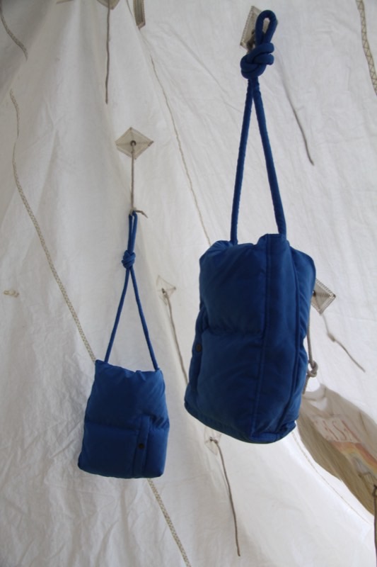 Two royal blue quilted bags hung by knotted rope straps against draped off-white sailcloth with paper kite tags.