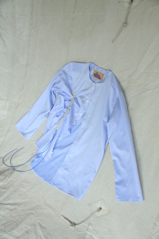 A collarless pale blue shirt with an asymmetric off-centre placket, a shirred vertical channel and trailing blue drawcords.