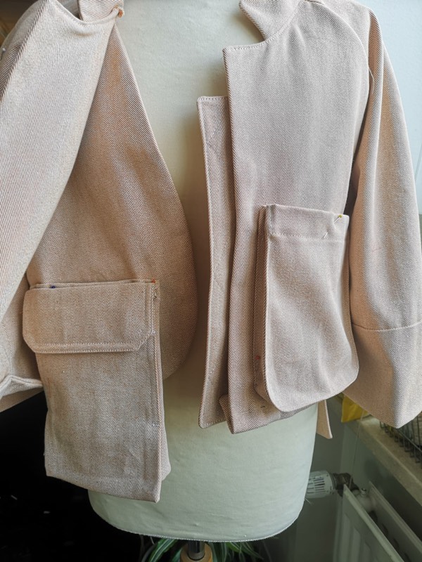 An undyed cotton twill jacket toile on a dress form in the studio, flap patch pockets still pinned in place.
