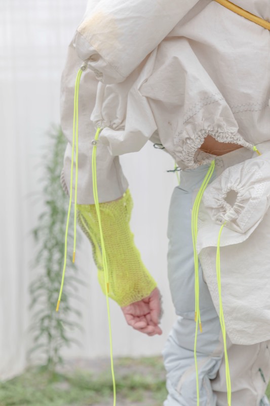 Close view of a cream padded nylon garment with drawcord eyelets trailing neon yellow cords, worn with a fluorescent open-knit fingerless sleeve.