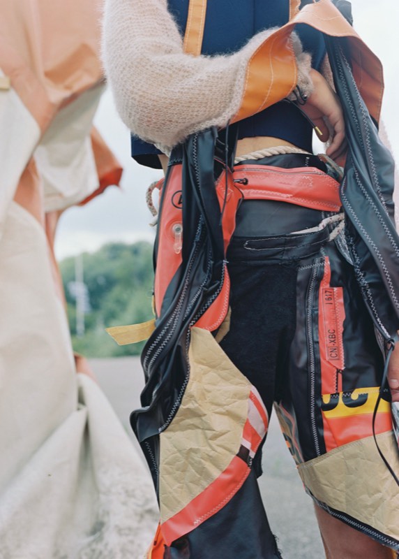 Rear detail of trousers patchworked from rescue-orange life-vest fabric, black rubber and tan panels over black denim, zigzag stitched, with a rope belt.
