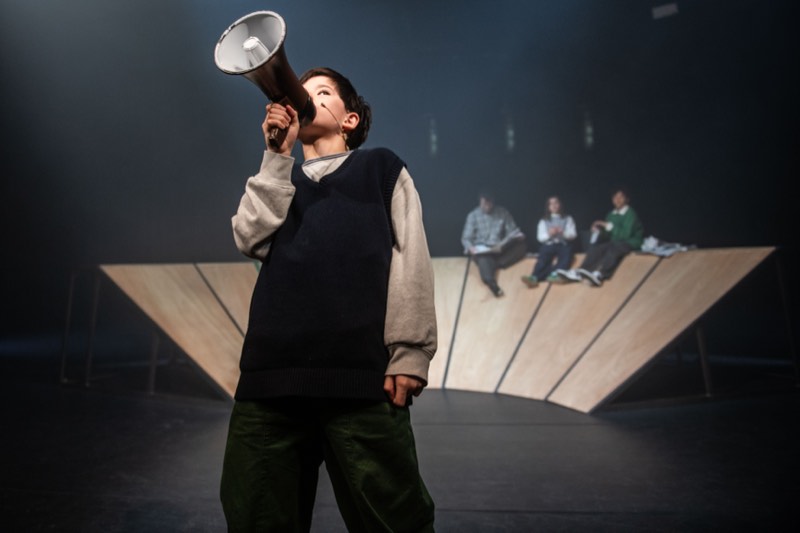 A backlit child downstage holds a megaphone, in a navy knitted tank over an oatmeal sweatshirt and wide olive corduroys.