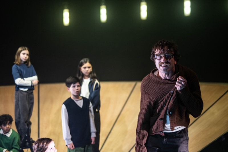 A man in a rust hand-knitted shawl-collar cardigan speaks mid-gesture, with four children behind him.