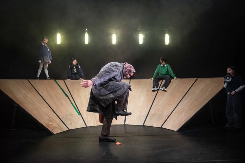 An adult in a long grey tweed overcoat bends double on a black stage while five children stand along a plywood set piece.