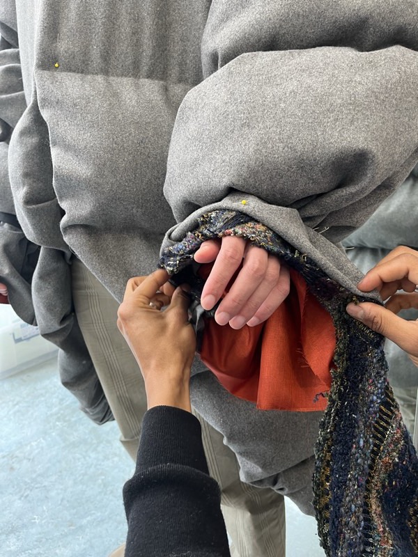 Two pairs of hands pin a hem where grey wool, a multicoloured boucle binding and raw frayed rust linen meet in one seam.