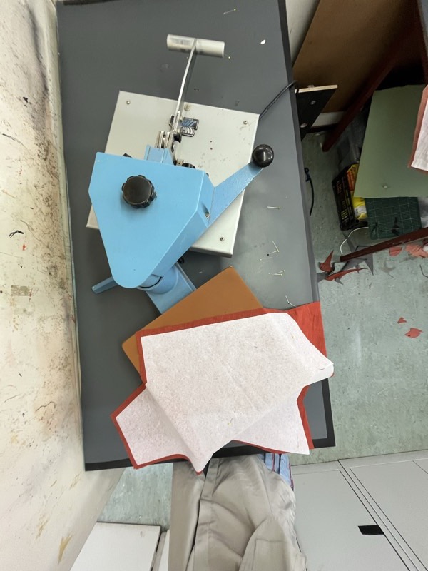 A small bench-top needle punch machine beside cut sheets of white and rust coloured felt on a workbench.