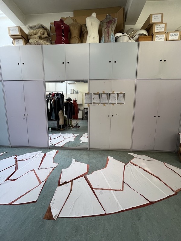 Large curved pattern panels edged in rust tape laid across an atelier floor, with a wall of storage and a costume rail behind.
