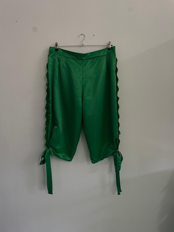 Emerald green satin cropped breeches on a wire hanger, side seams edged with knife-pleated ruffle trim and gathered onto ribbon ties.