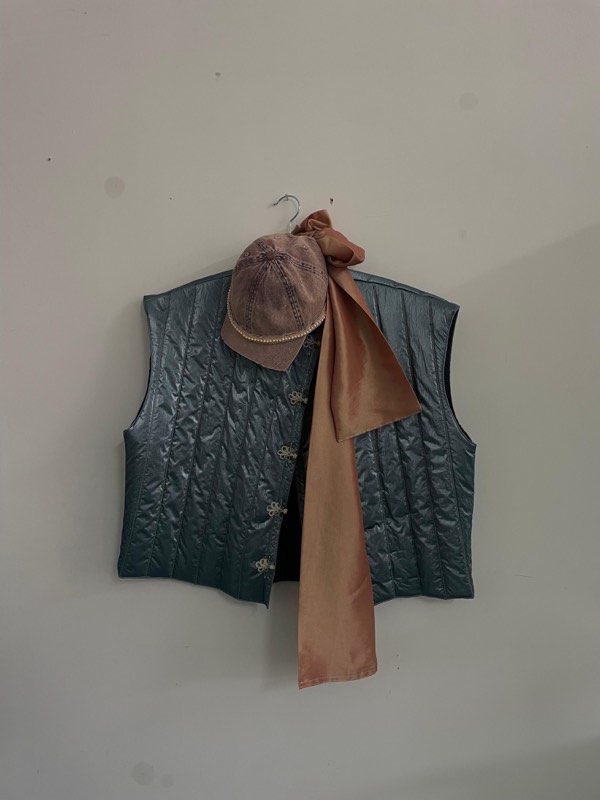A petrol teal quilted waistcoat with filigree metal buttons hangs with a mauve suede flat cap and a long copper satin sash.
