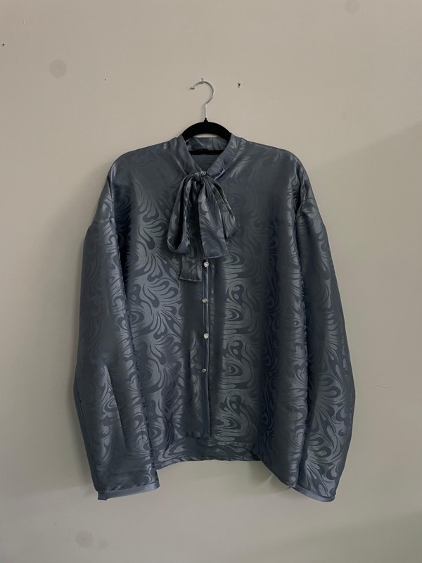 A boxy grey-blue jacquard shirt with a large paisley figure, pearl buttons and an attached pussy-bow cravat, on a black hanger.