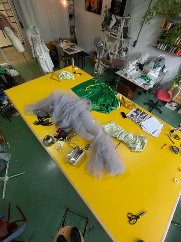 A wide view of the workroom: a bright yellow cutting table heaped with grey tulle, emerald satin and shears, with sewing machines behind.