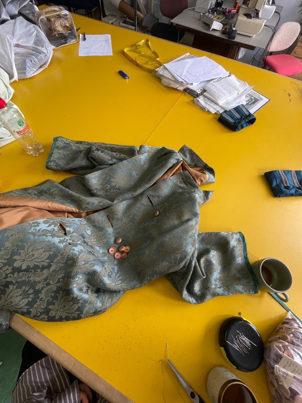 A pale blue-green brocade tailcoat lies open on a yellow worktable with copper covered buttons, braid-trimmed teal cuffs and handwritten measurements.