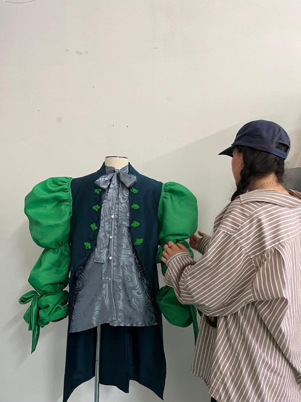 The maker adjusts a navy waistcoat with appliqued leaf motifs and large emerald satin puff sleeves on a dress form.