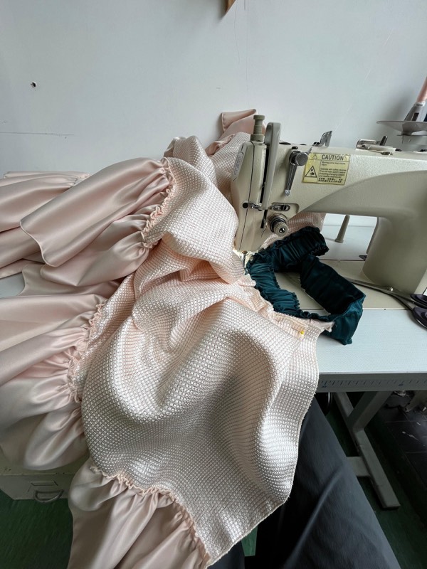 The blush ensemble under the needle of an industrial machine, with pale pink satin puff sleeves and a teal ruched velvet waistband.