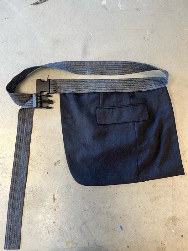 A wrap apron panel cut from navy suit wool with a welt pocket, hanging from a pinstriped fabric belt with a black side-release buckle.