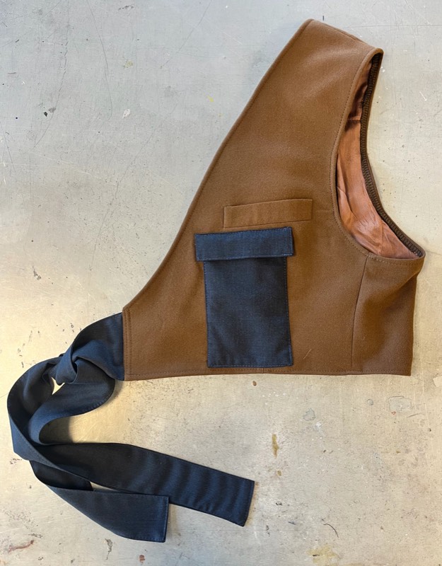A cropped tobacco-brown canvas waistcoat with a navy flap pocket, leather-bound armhole and long navy sash ties, laid flat on a concrete floor.