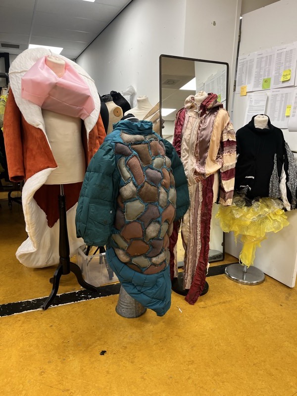 A teal quilted puffer jacket appliqued all over with irregular polygonal patches of suede, felt and hessian, on a rail of costumes in the wardrobe store.