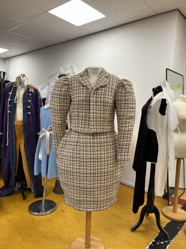 A brown and cream houndstooth suit on a wooden dress form, the cropped jacket with strongly gathered puff sleeve heads.