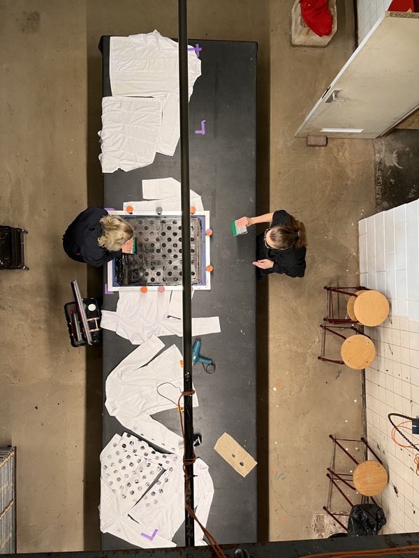 Two people screen-print black dots onto white workwear jackets laid out on a long print table, seen from above.