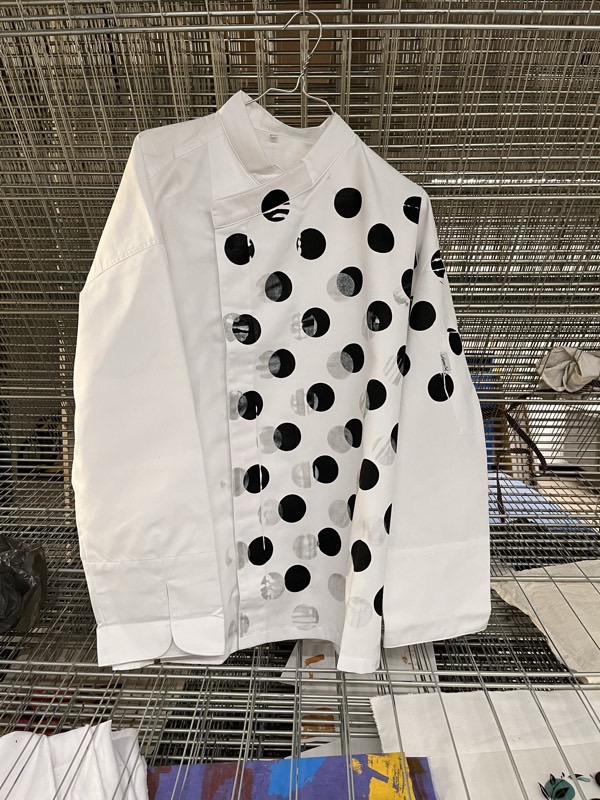 A white double-breasted workwear jacket on a wire drying rack, printed with a diagonal grid of black and ghosted grey dots.
