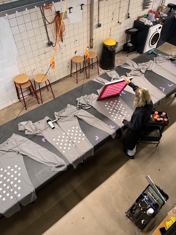 Seen from above, the designer lifts a hot pink screen frame off engineer-striped trousers laid along a black print table.
