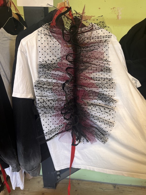 A white T-shirt on a rack with a constructed spine of black spotted and magenta tulle ruffles down the centre back and loose red ribbon ties.
