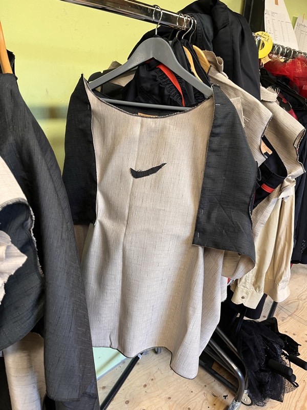 An unlined oatmeal slub bodice hanging on a rolling rack with a charcoal panel pinned over one shoulder.