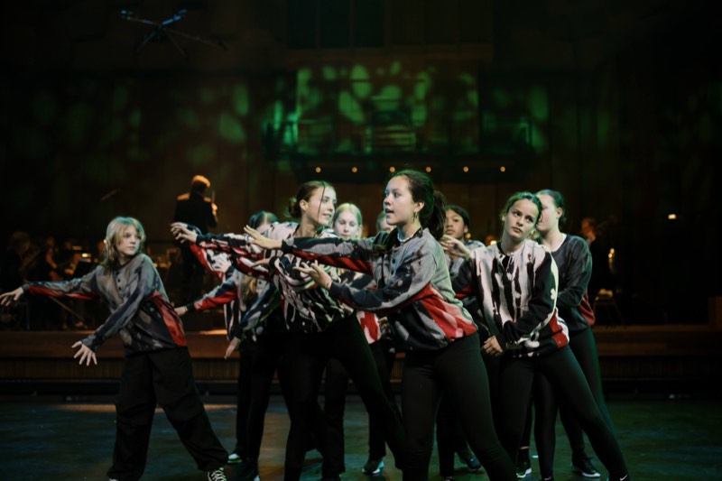 Nine young performers cluster mid-movement in bomber jackets printed with a brushed grey, white and blood-red camouflage.