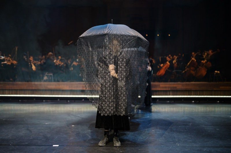 A single figure centre stage under a transparent umbrella skinned in black floral lace, in a matching lace coat over a velvet maxi skirt.