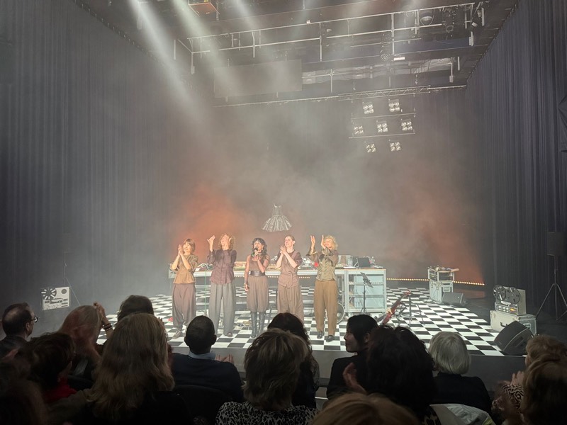 Curtain call seen from the audience, five performers bowing in layered taupe separates with the silver bell skirt suspended above the stage.