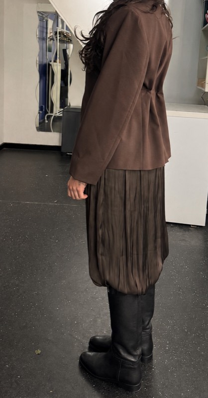 Fitting photograph from behind of a brown wool blazer with a shaped waist over a pleated chiffon skirt and black riding boots.