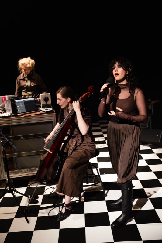 Three performers in brown: a sheer mesh layer over a slip and a draped balloon skirt, a wide pinafore trouser, and an olive utility shirt.
