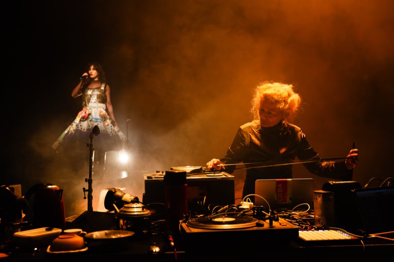 A performer draws a bow across a table of turntables in orange smoke while a singer upstage wears a stiff metallic bell skirt of collaged panels.