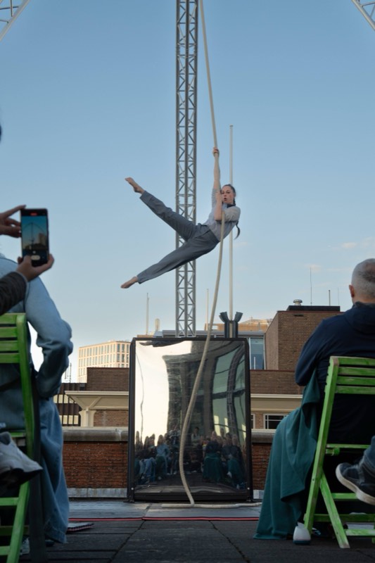 An aerialist holds a vertical rope in a horizontal split above a rooftop audience, in grey wide-leg trousers and a panelled grey shirt.