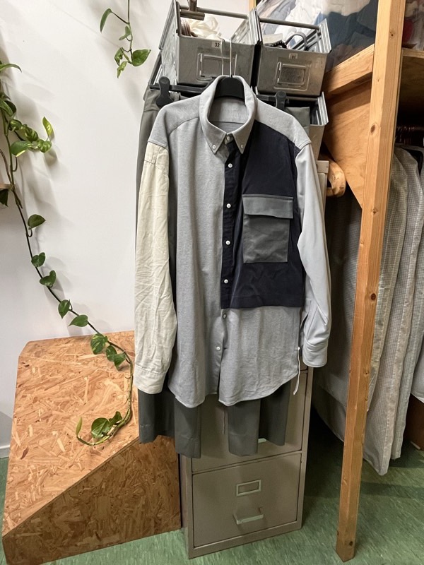 A grey jersey shirt dress with contrasting ecru sleeves hangs on a studio rail under a navy panelled vest with a large flap pocket.