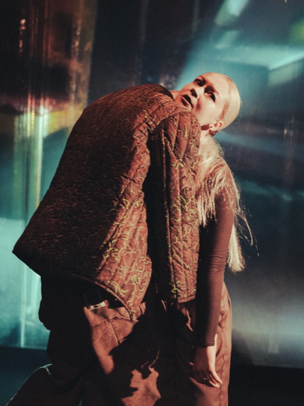 A dancer arches backwards in a brown quilted jacket stitched with looping lime green quilting lines, under teal and amber stage light.