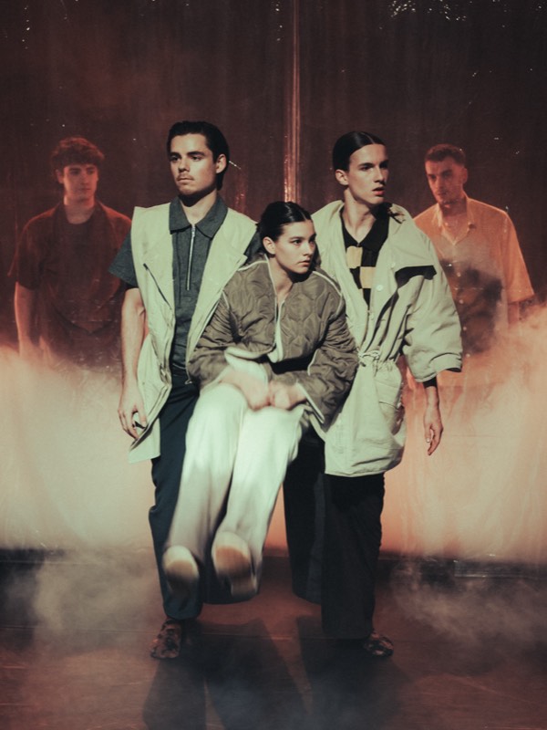 Five performers advance through haze in sand and olive outerwear, one lifted between them in a quilted olive jacket and wide ivory trousers.