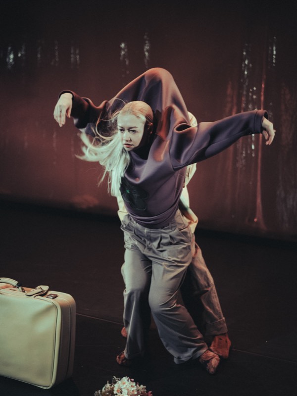 A dancer swings forward mid-movement, arms outstretched, in an oversized aubergine sweatshirt torn at the shoulder and cuffed grey trousers.