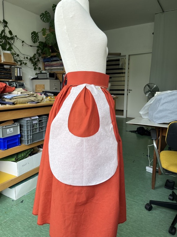 A full rust coloured skirt on a dress form with a white interfacing apron prototype pinned over it, cut with a keyhole opening.