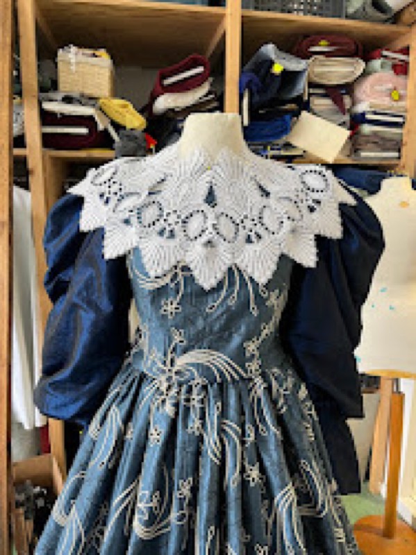 A blue embroidered period bodice with large puffed navy sleeves under a wide cream cutwork lace collar, on a dress form.